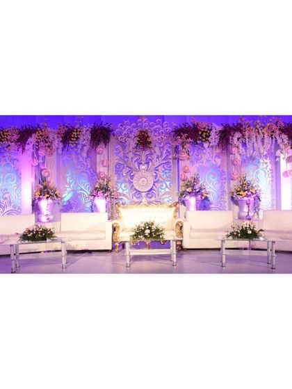 An exceptionally detailed and luxurious reception stage. The intricate white and gold carved backdrop, illuminated by soft purple lighting and adorned with flowers, provides a truly regal setting for the couple.