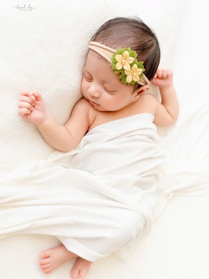 A newborn with a green floral headband sleeps peacefully, wrapped in a simple white cloth that emphasizes their natural beauty.