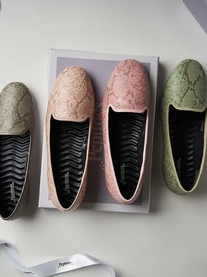 A top-down flat lay for Pepitoes, showcasing their snakeskin-textured loafers. The composition with the box and ribbon gives it a "new shoes" feel, and the soft lighting is perfect for showing off the pastel colors.
