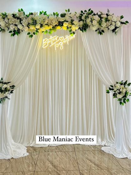 A grand and elegant backdrop perfect for a Roka or reception stage. The design features a wide wall of white drapes, a floral runner across the top, and a 'Better Together' neon sign for a clean and classic look.