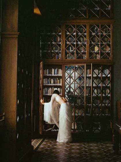 Exploring the library, the sheer white saree interacts beautifully with the dark wood and natural light, adding to the romantic, scholarly atmosphere of the scene.