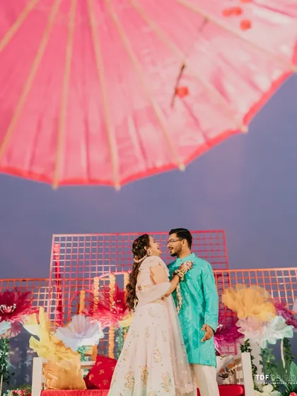 A creative shot of the couple under a bright pink umbrella, set against the vibrant floral backdrop of their Haldi carnival. I love finding unique angles to capture their love story.