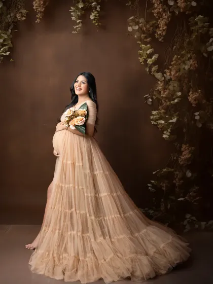 An earthy and romantic portrait. The mother-to-be wears a flowing, tiered nude gown, standing against a brown backdrop with cascading foliage.
