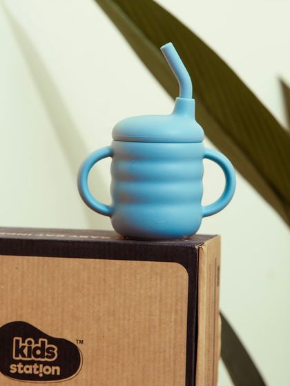 A close-up of a blue sippy cup resting on its branded box. This is a clean product shot that highlights the design and branding.