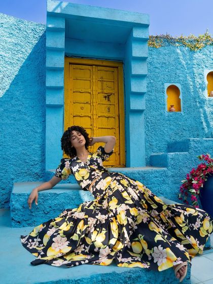 A relaxed, seated pose from the Tangerine Tiger campaign, showcasing a floral two-piece set against a vibrant blue wall.