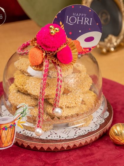 Celebrate the warmth of Lohri with this elegant glass cloche filled with homemade til and jaggery cookies. The festive decorations add a perfect touch of tradition.