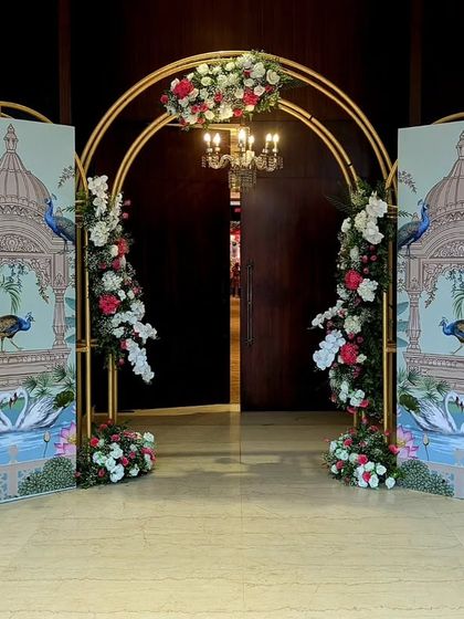 A unique entrance featuring custom panels with traditional Indian paintings of peacocks and palaces. The golden arches and floral arrangements blend artistry with classic wedding decor for a truly bespoke welcome.