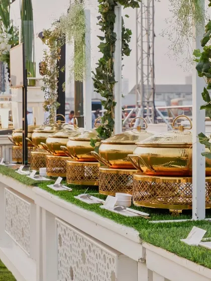 A row of gleaming golden chafing dishes on a pristine white buffet counter. This setup, adorned with fresh greenery, is a picture of elegance and readiness.