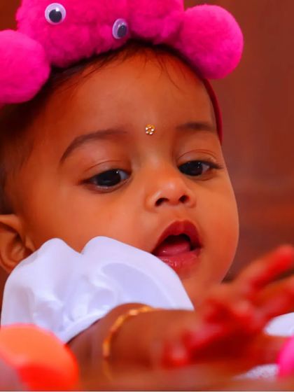 A playful close-up of a baby girl with a fun, fuzzy pink headband. This shot captures her curiosity as she reaches for something just out of frame.