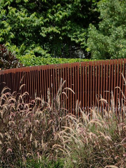A curved, rust-colored Corten steel screen provides a semi-private boundary in a garden. The screen's vertical slats and gentle curve interact beautifully with the soft textures of the surrounding ornamental grasses.
