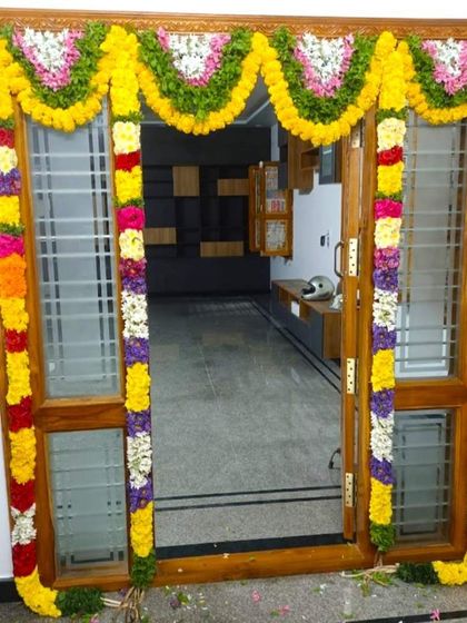 An entrance door flower decoration for a housewarming. The wooden door with glass panels is framed by colorful traditional flower garlands.