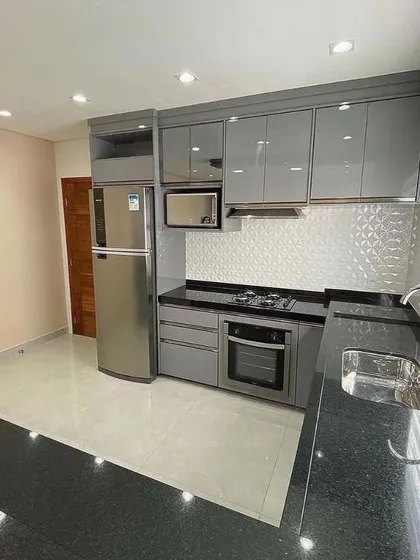 A view of a compact kitchen layout with grey high-gloss cabinets and a 3D-textured white tile backsplash. The appliances, including the fridge, microwave, and oven, are all neatly integrated.