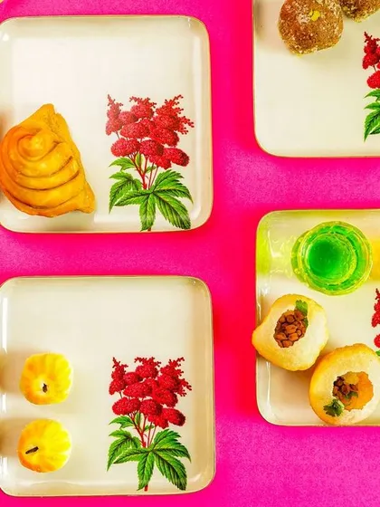 A vibrant flatlay of square, floral-patterned plates with Indian sweets, set against a bright pink background.