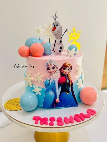 Frozen-themed cakes are always a favorite. This design features Elsa, Anna, and a cheerful Olaf, surrounded by chocolate spheres and snowflakes.