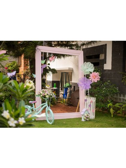 A whimsical photo booth setup in a garden, featuring a large pink-framed mirror, a vintage-style bicycle, and colorful paper flowers.