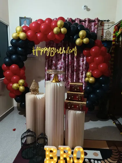 A classic balloon arch is a timeless choice for any celebration. This setup for a brother's birthday uses a bold red, black, and gold color combination, along with light-up letters and pedestals for gifts and cake.
