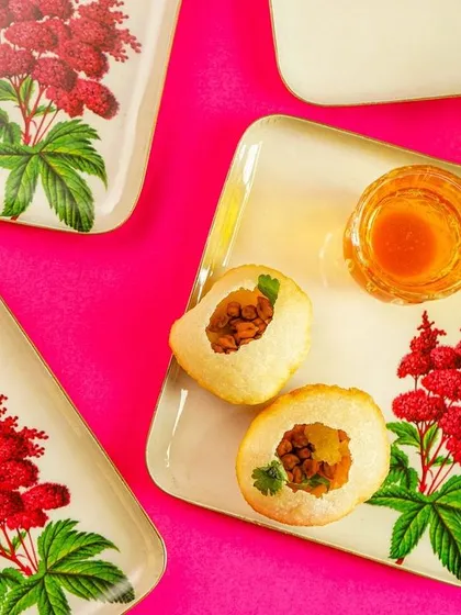 A vibrant flatlay of square floral plates with Indian sweets, shot against a bright pink background.