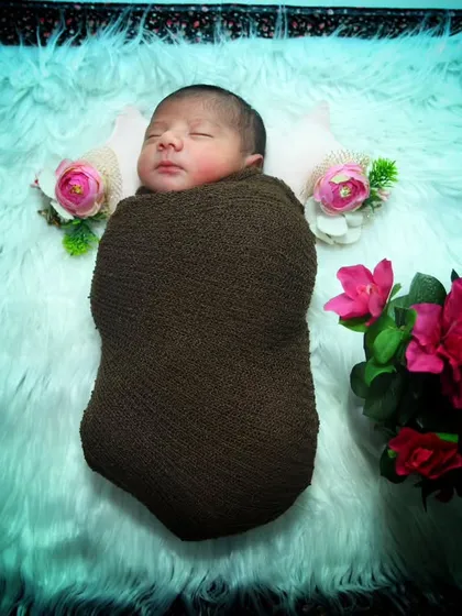 A classic newborn pose. This baby is sleeping soundly on a white fur rug, wrapped in a brown knit swaddle and adorned with small flowers.