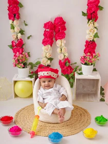 A smiling baby enjoying the colorful Holi setup from the comfort of his little chair.