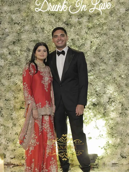 The happy couple posing at the floral photo wall. The bride's vibrant red outfit provides a stunning contrast against the white backdrop.