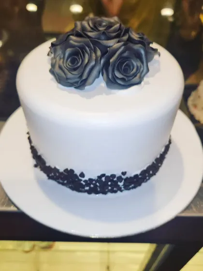 A striking and minimalist cake design. The clean white fondant is contrasted with a cluster of black-dusted roses on top and a border of chocolate sprinkles, making for a modern and sophisticated statement.