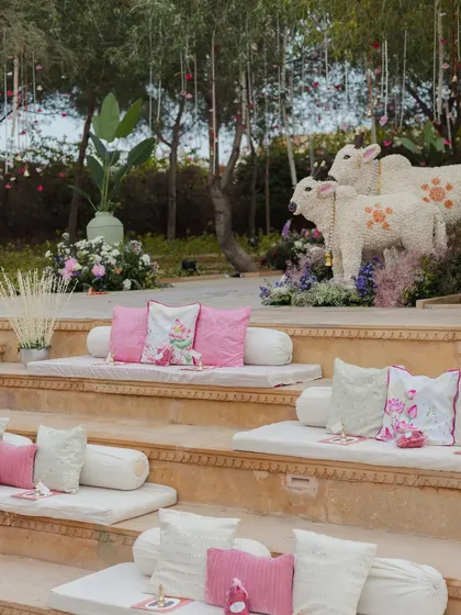 Serene baithak seating on stone steps, accented with pink cushions and floral cow installations, creating a tranquil and traditional space for guests.