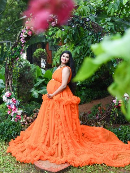 A vibrant portrait of a mom-to-be in an orange gown, framed by a rustic arch and lush green foliage.