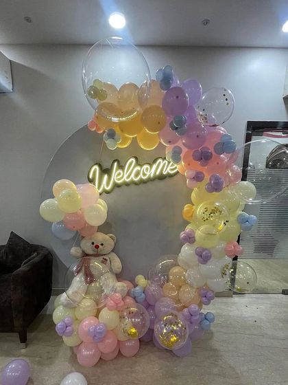 This pastel-themed welcome decoration features a circular backdrop with a neon sign, a cute teddy bear, and an organic balloon garland. It's a perfect, gentle theme for a newborn.