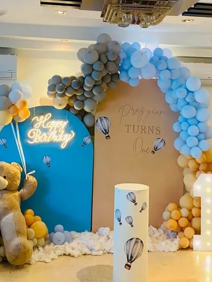 A whimsical 'hot air balloon and teddy bear' theme for a first birthday party. This setup includes a custom arched backdrop, a light-up number '1', and a life-sized teddy bear, all tied together with a soft blue, grey, and yellow color palette for a picture-perfect celebration.
