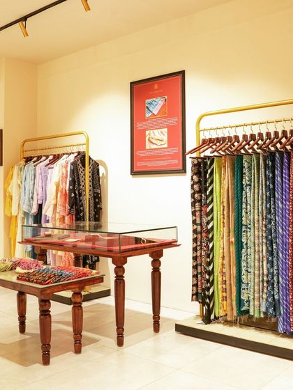 The clothing rack and display area in the 'House of Vaarasa' showroom. We used minimalist brass-finished racks and hand-carved wooden tables to let the colorful garments stand out, creating a boutique shopping experience.