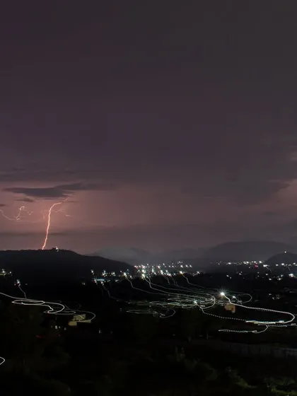 A long-exposure shot captures a lightning strike over Pune, combined with the light trails of vehicles on a winding road.
