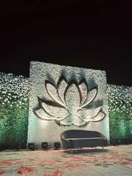 A minimalist yet striking stage design featuring a large 3D lotus cutout against a wall of white flowers. The clean lines and focused lighting create a serene and elegant photo backdrop.