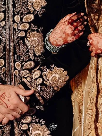 A close-up on the details, showing the bride's henna-adorned hand on the groom's intricately embroidered sleeve.