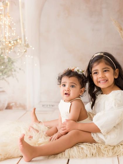 Two sisters sit together in a beautifully styled boho-chic studio setup. The older sister holds the younger one, showcasing their sweet bond.