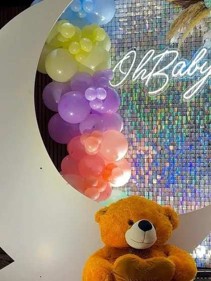 A close-up of the crescent moon prop, filled with pastel rainbow balloons and set against a silver shimmer wall. The "Oh Baby" neon sign and a cute teddy bear complete this perfect photo opportunity.