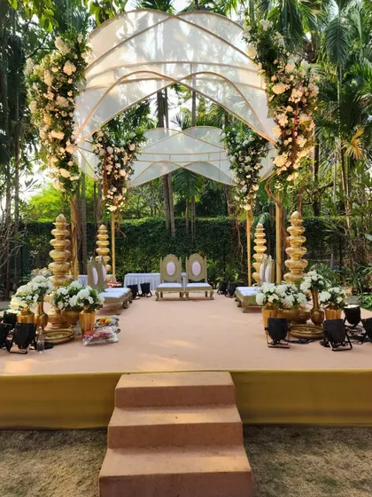 This unique mandap design for an outdoor wedding features a modern arched roof structure draped in sheer fabric and adorned with clusters of white flowers, creating an airy and elegant space.