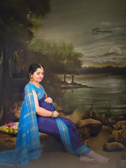 A serene studio portrait of a mother-to-be in a blue nauvari saree, seated against a painted landscape backdrop.