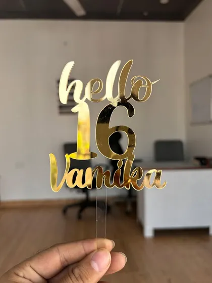 Holding the "Hello 16 Vamika" topper to demonstrate its size and how the gold mirror finish catches the light.