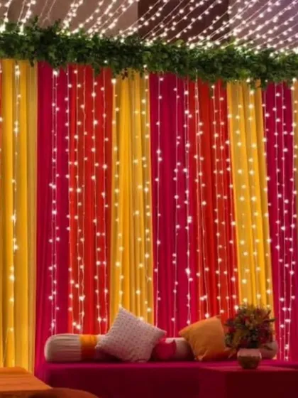 A cozy seating area with a backdrop of colorful drapes and fairy lights. The pink and yellow fabric creates a warm and inviting atmosphere for a Sangeet or Mehandi night.