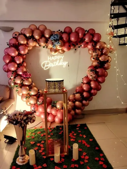 An indoor birthday or anniversary setup with a large heart-shaped balloon arch in metallic rose gold. A "Happy Birthday" neon sign and a cake stand are placed in the center, with rose petals on the floor for a touch of romance.