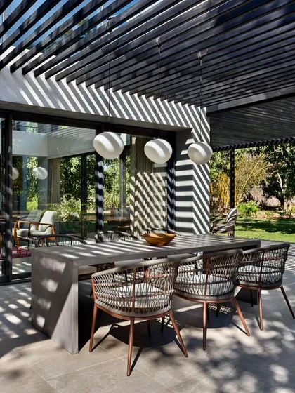 The outdoor dining table is set under a pergola, where the slatted roof creates a dynamic pattern of sunlight on the surfaces below. This space is designed for enjoying meals in the fresh air.