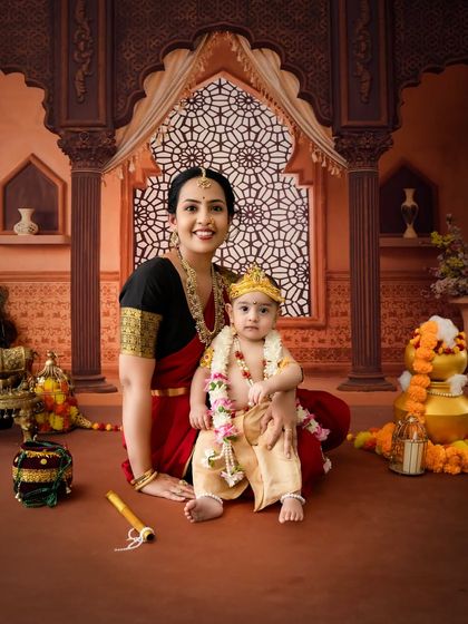 A classic portrait of a mother and her son in a traditional Janmashtami setup, seated before an ornate window backdrop.