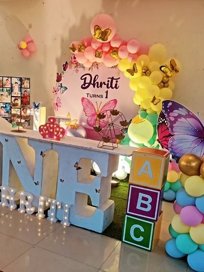 A large-scale butterfly-themed first birthday. The setup includes a 'ONE' marquee table, a photo collage board, and colorful balloon columns.