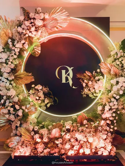 A personalized photo backdrop for the couple, featuring their monogram inside a neon-lit ring and surrounded by an explosion of white florals and golden palm leaves.