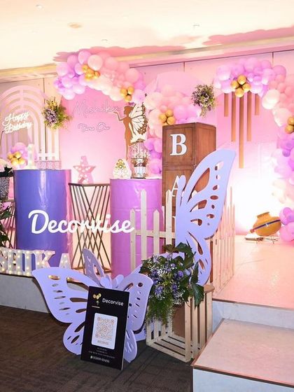 This wide view shows the complete butterfly-themed setup for Mishika's party. The stage includes a main backdrop with a balloon arch, a milestone board detailing her first year, and decorative props like wooden crates and a QR code stand for guests.