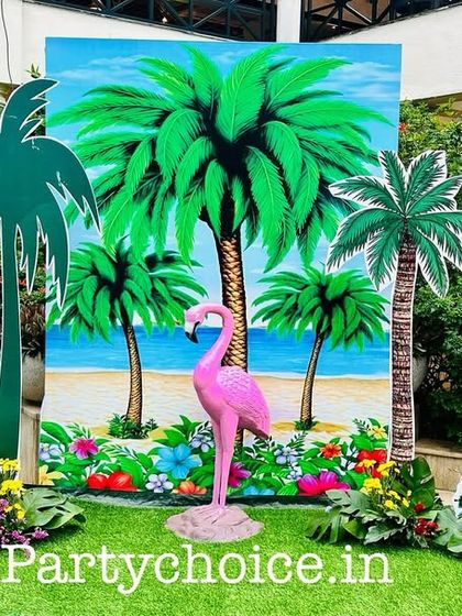 A beach-themed photo booth set up on the grass, featuring a painted backdrop, palm tree cutouts, and tropical bird props.