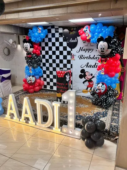 A classic Mickey Mouse first birthday party, featuring a checkered backdrop, balloon columns with Mickey foils, and large light-up letters for the name 'AADI'.