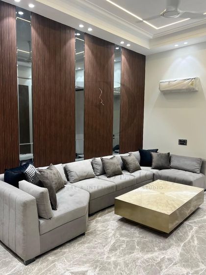This L-shaped sectional sofa in a soft grey fabric provides ample seating, paired with a striking brass-finished center table. The back wall features a modern design of wood veneer and mirror panels, adding depth to the room.