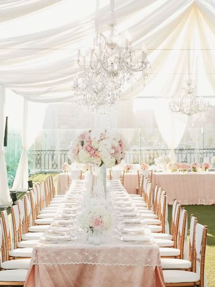 An elegant long-table dining setup for a wedding reception. The all-white decor with grand chandeliers creates a sophisticated and luxurious atmosphere.