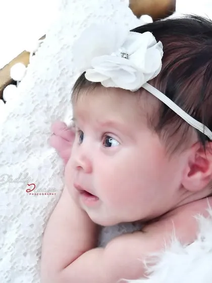Capturing a beautiful moment with an awake newborn, looking curiously to the side. The baby is wearing a delicate white flower headband and is nestled in soft white fur.